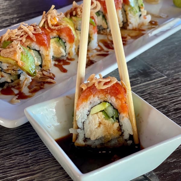 Chopsticks holding a sushi roll with avocado, red topping, and crispy fried onions, dipping into soy sauce.