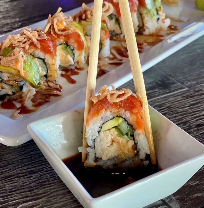 Chopsticks holding a sushi roll with avocado, red topping, and crispy fried onions, dipping into soy sauce.