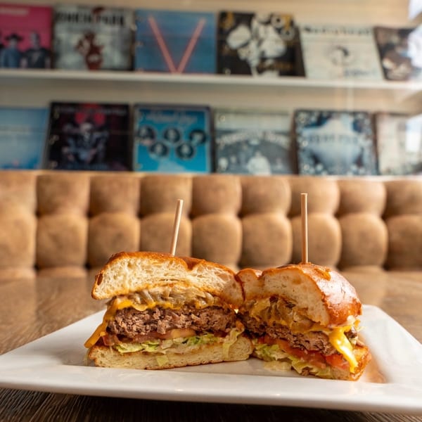 A burger cut in half on a white plate, with a blurred background of record albums.