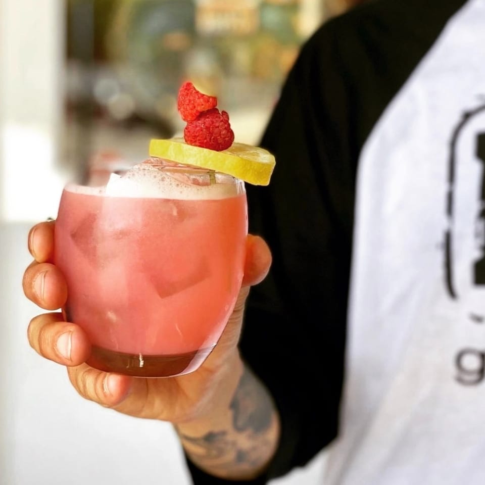 Hand holding a pink cocktail garnished with lemon and raspberries, against a blurred background.