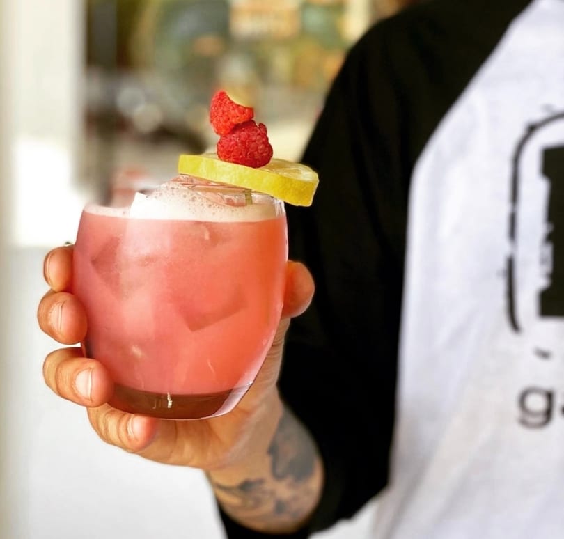 Hand holding a pink cocktail garnished with lemon and raspberries, against a blurred background.