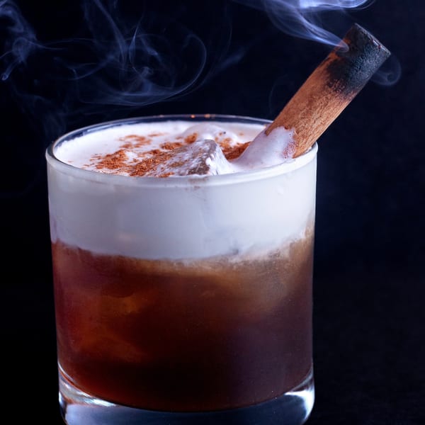 A smoked cocktail with a cinnamon stick, foamy top, and ice, against a dark background.