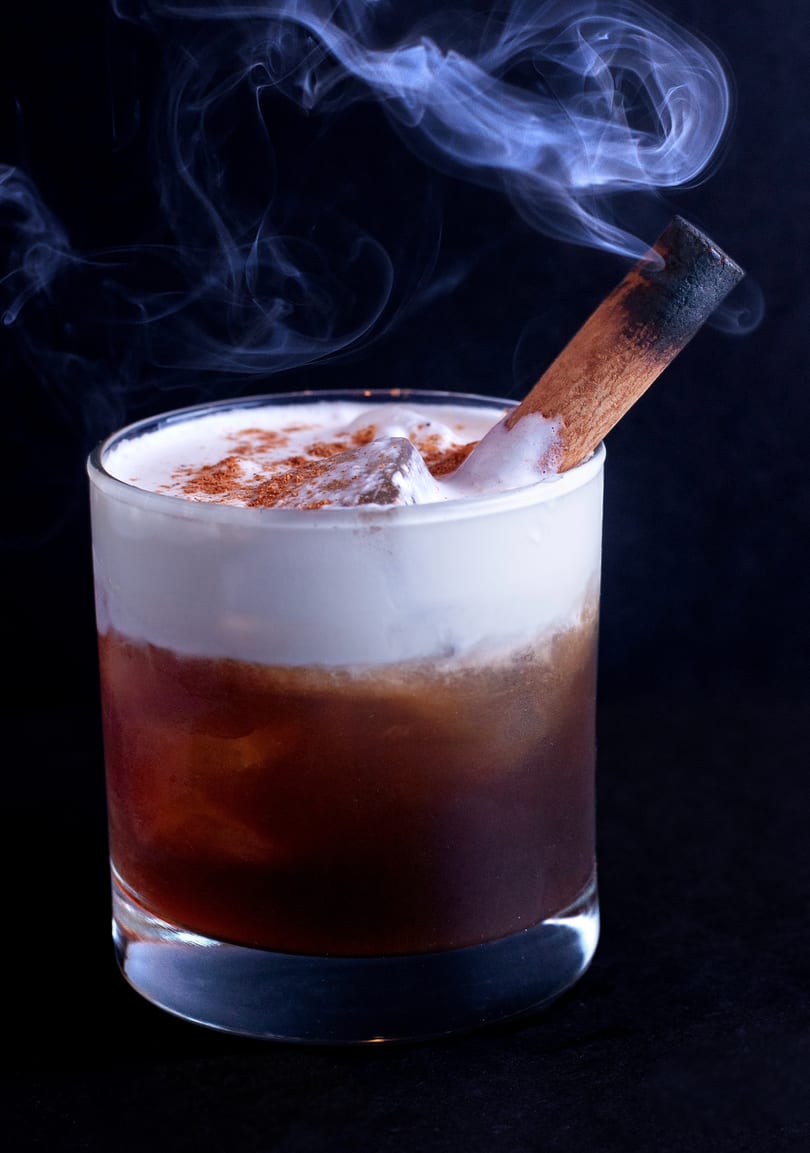 A smoked cocktail with a cinnamon stick, foamy top, and ice, against a dark background.