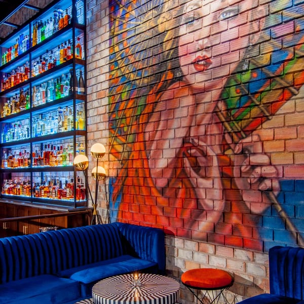 A vibrant bar interior with blue velvet seating, striped tables, and a colorful mural on a brick wall.