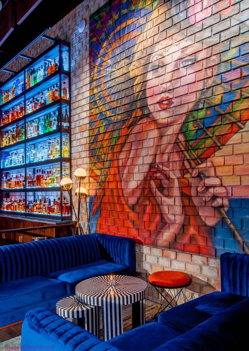 A vibrant bar interior with blue velvet seating, striped tables, and a colorful mural on a brick wall.