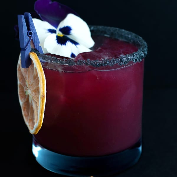 A dark red cocktail with a black sugar rim, garnished with a dried orange slice and a pansy.