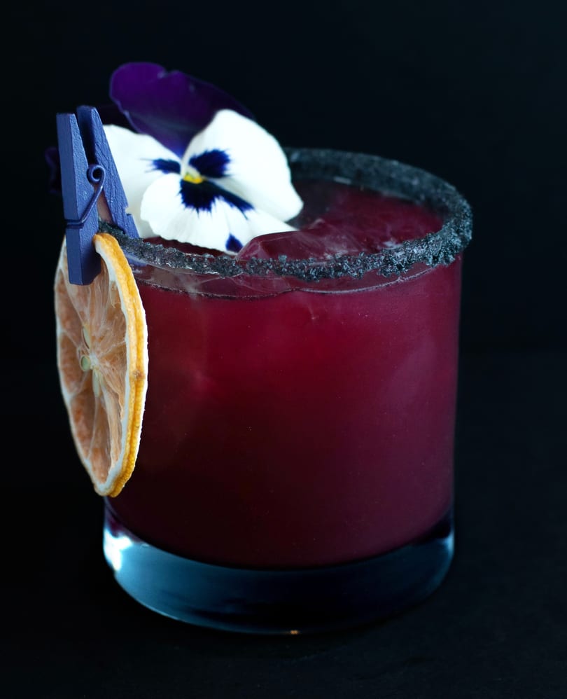 A dark red cocktail with a black sugar rim, garnished with a dried orange slice and a pansy.