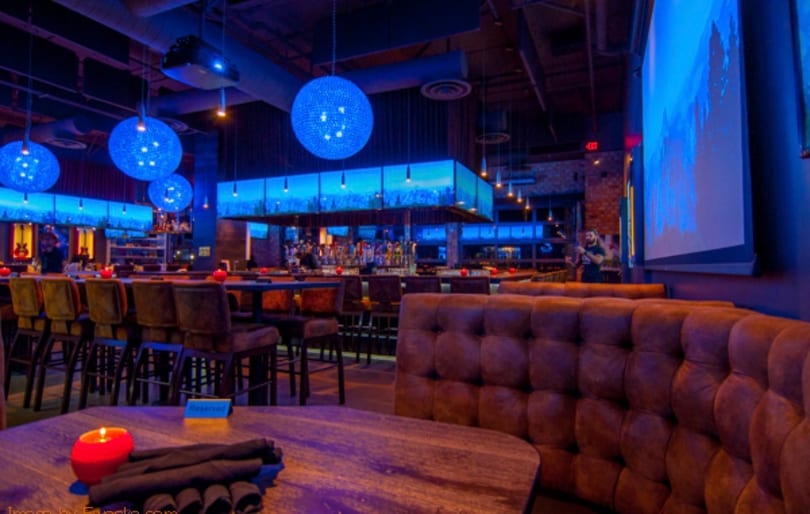 Restaurant interior with blue lighting, large round chandeliers, a bar, and brown booth seating.