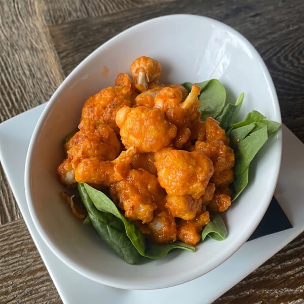A white bowl filled with orange buffalo cauliflower bites and fresh spinach on a wooden table.