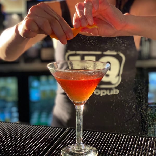 Bartender squeezing an orange peel over a red cocktail in a martini glass at a bar.