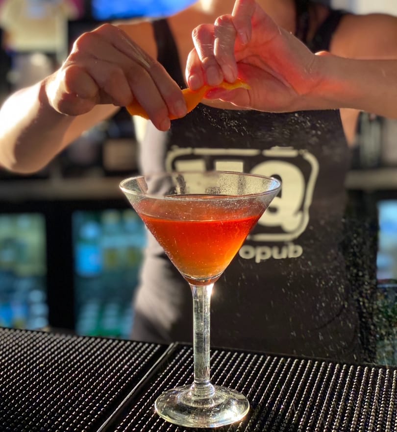 Bartender squeezing an orange peel over a red cocktail in a martini glass at a bar.