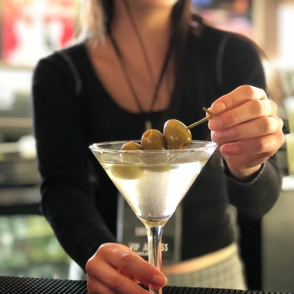 A bartender garnishes a martini with olives, holding the glass on a bar mat.