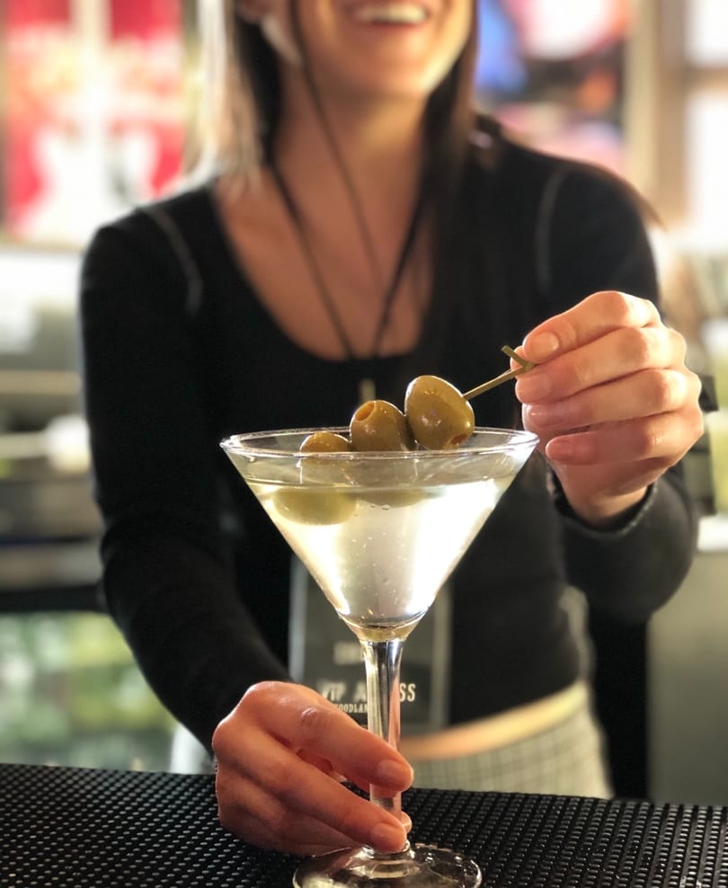 A bartender garnishes a martini with olives, holding the glass on a bar mat.
