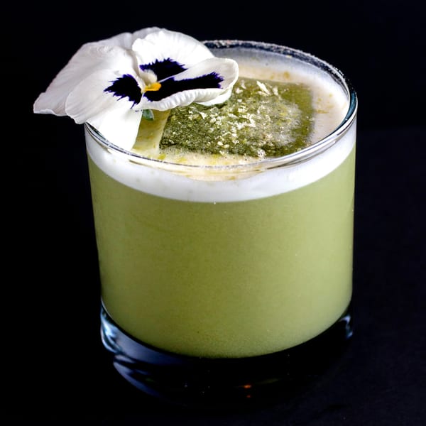 Green cocktail in a glass with a white and purple pansy garnish, against a dark background.