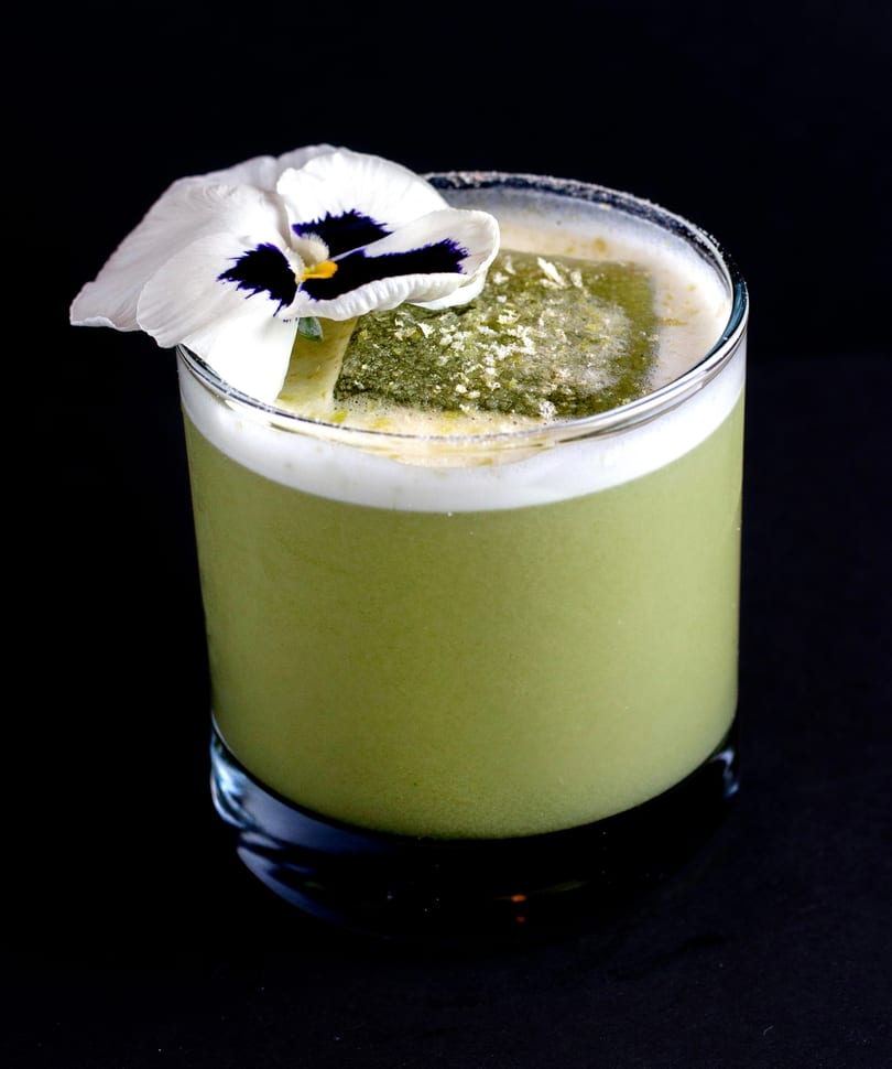 Green cocktail in a glass with a white and purple pansy garnish, against a dark background.